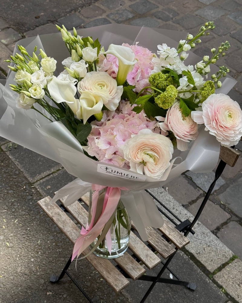 Pastellfarbener Strauß mit Ranunkeln, Calla und rosa Hortensie in mattweißem Papier mit LUMIÈRE-Etikett, in einer Glasvase auf einem Bistrostuhl aus Holz