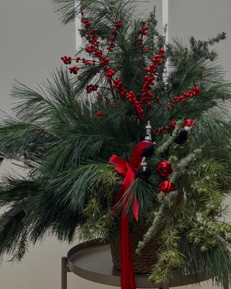 Weihnachtsgesteck aus Kiefern- und Tannenzweigen mit roten Beeren, roten und schwarzen Kugeln und roter Schleife in einem geflochtenen Korb